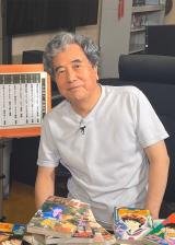 A candid color photograph of manga artist Yū Koyama for an Azumi manga review. He is seen in a relaxed setting, wearing a blue button-down shirt and glasses, smiling warmly. He appears to be in an office or studio environment with books and papers in the background.