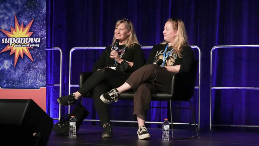 Veronica Taylor Interview: The Voice of Ash on Dubbing, Lip Flap and 25 Years of Pokémon 8 veronica and rena taylor sitting in black chairs on a stage during a panel at Supanova Comic Con & Gaming. The woman on the left, voice actress Veronica Taylor, is holding a microphone and smiling as she speaks. She is wearing a black long-sleeved top, dark pants, and black sneakers. The woman on the right is listening attentively, wearing a black graphic tee, brown pants, and black and white platform sneakers. Behind them is a dark blue curtain and to their left is a banner featuring the Supanova Comic Con & Gaming logo.